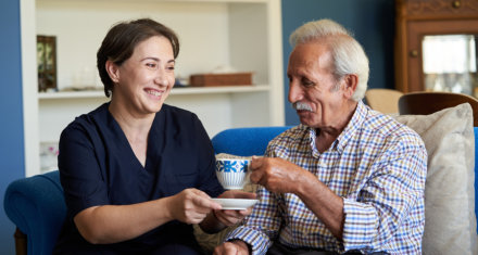 female caregiver smiling at an elderly man drinking tea 